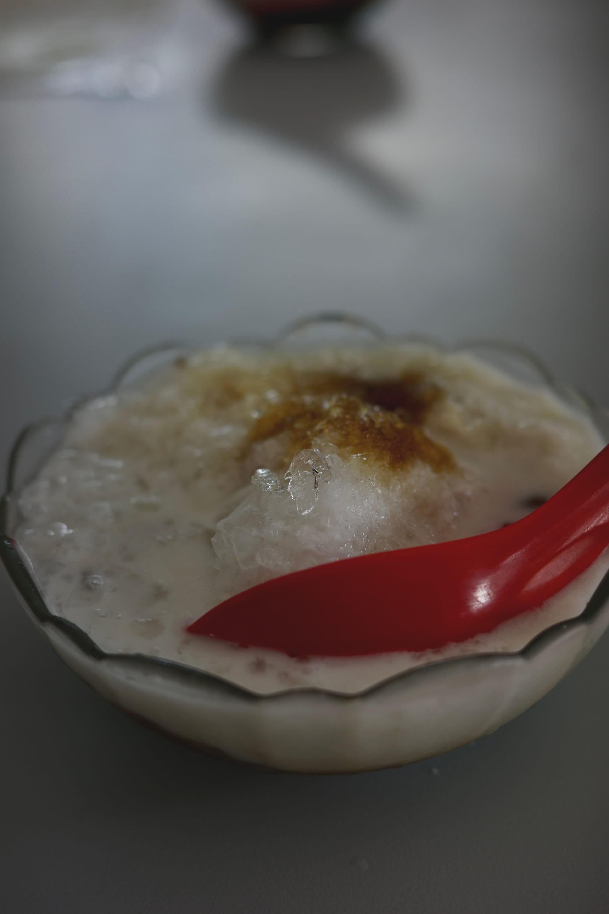 A glass bowl of shaved ice with a brown patch of palm sugar. A red spoon is stuck into the bowl