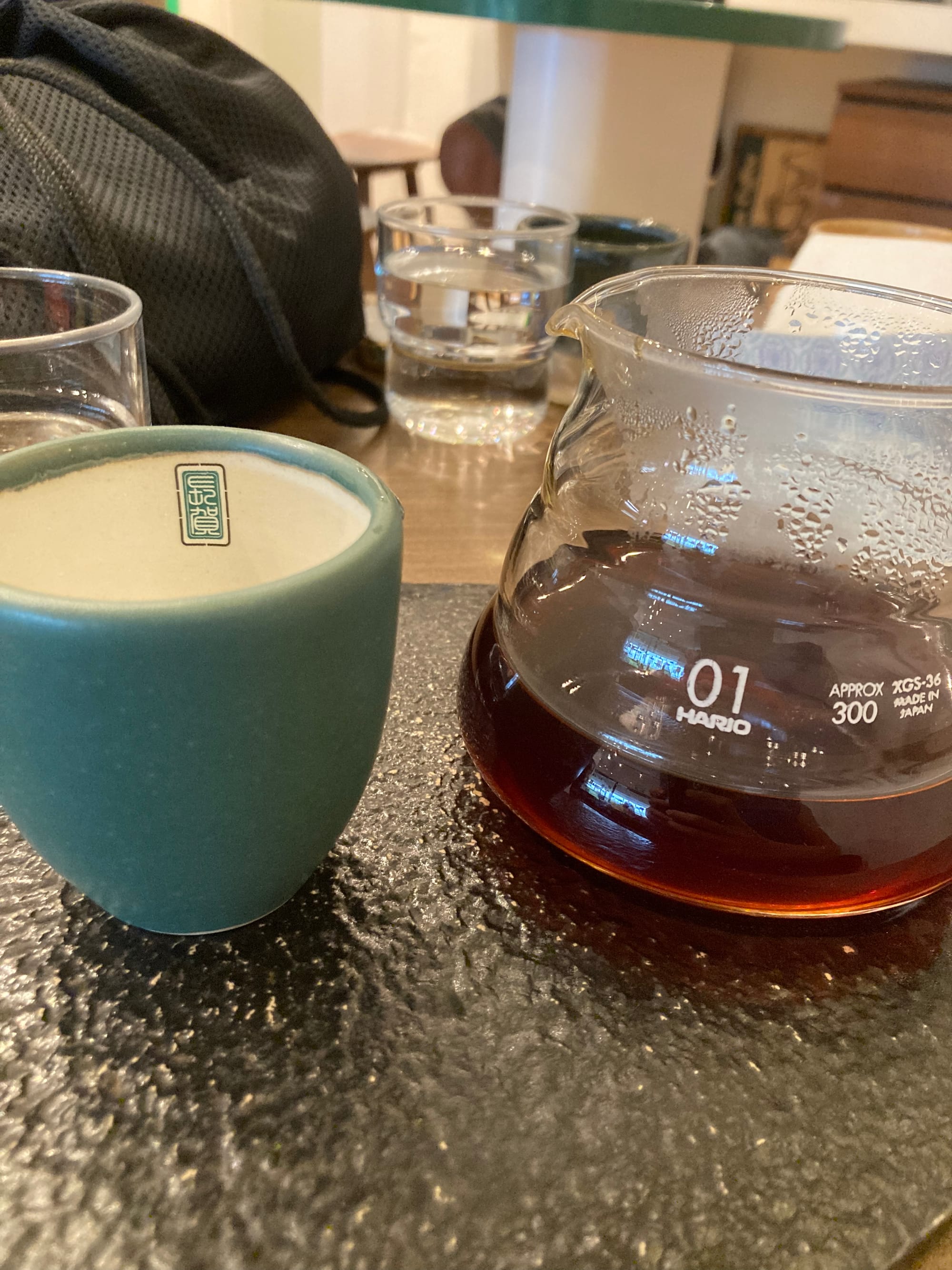 A glass pitcher of coffee brewed by hand next to a pale teal ceramic cup