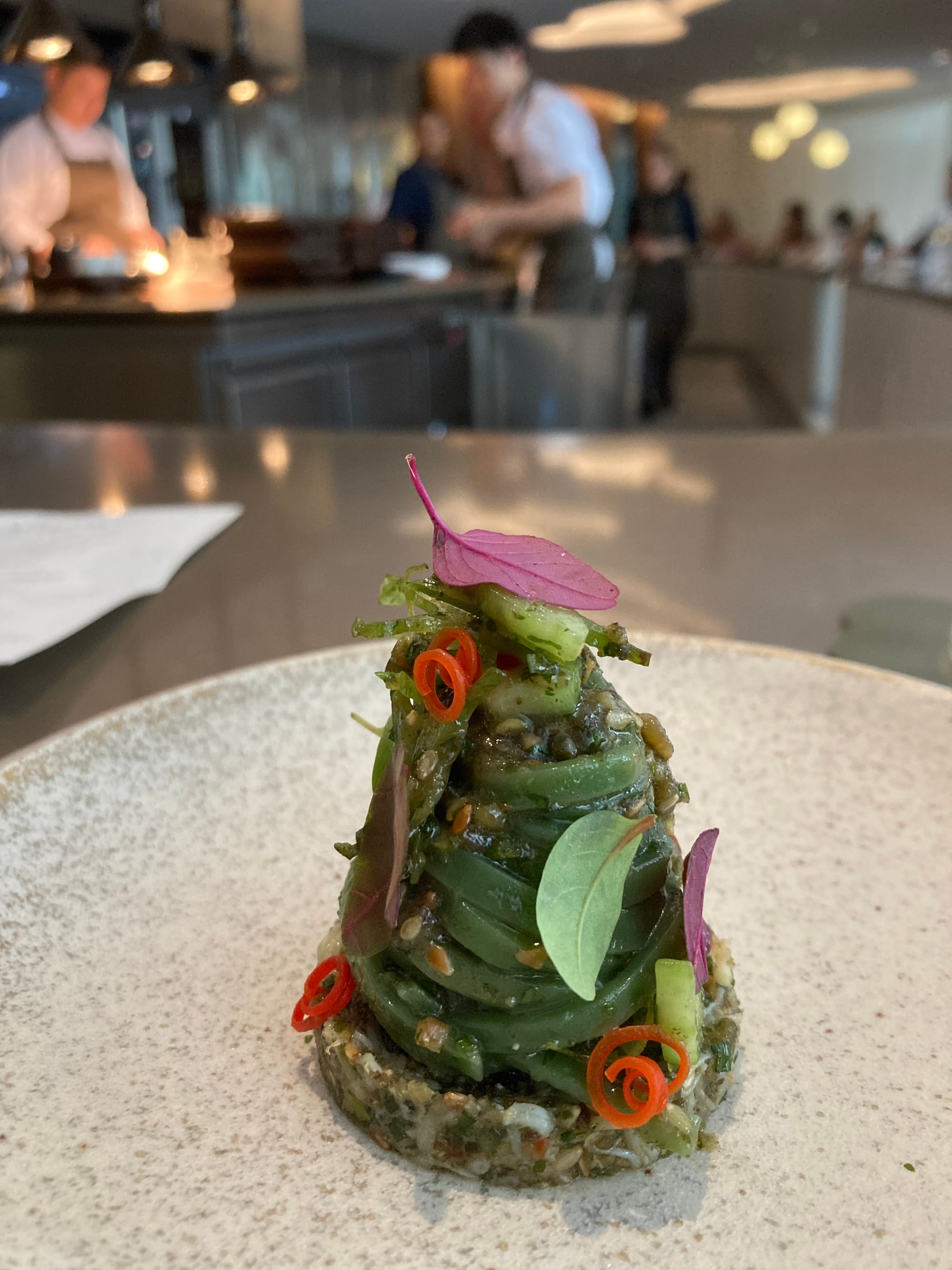 In the foreground is a neat pile of green noodles. In the background staff work in an open kitchen.