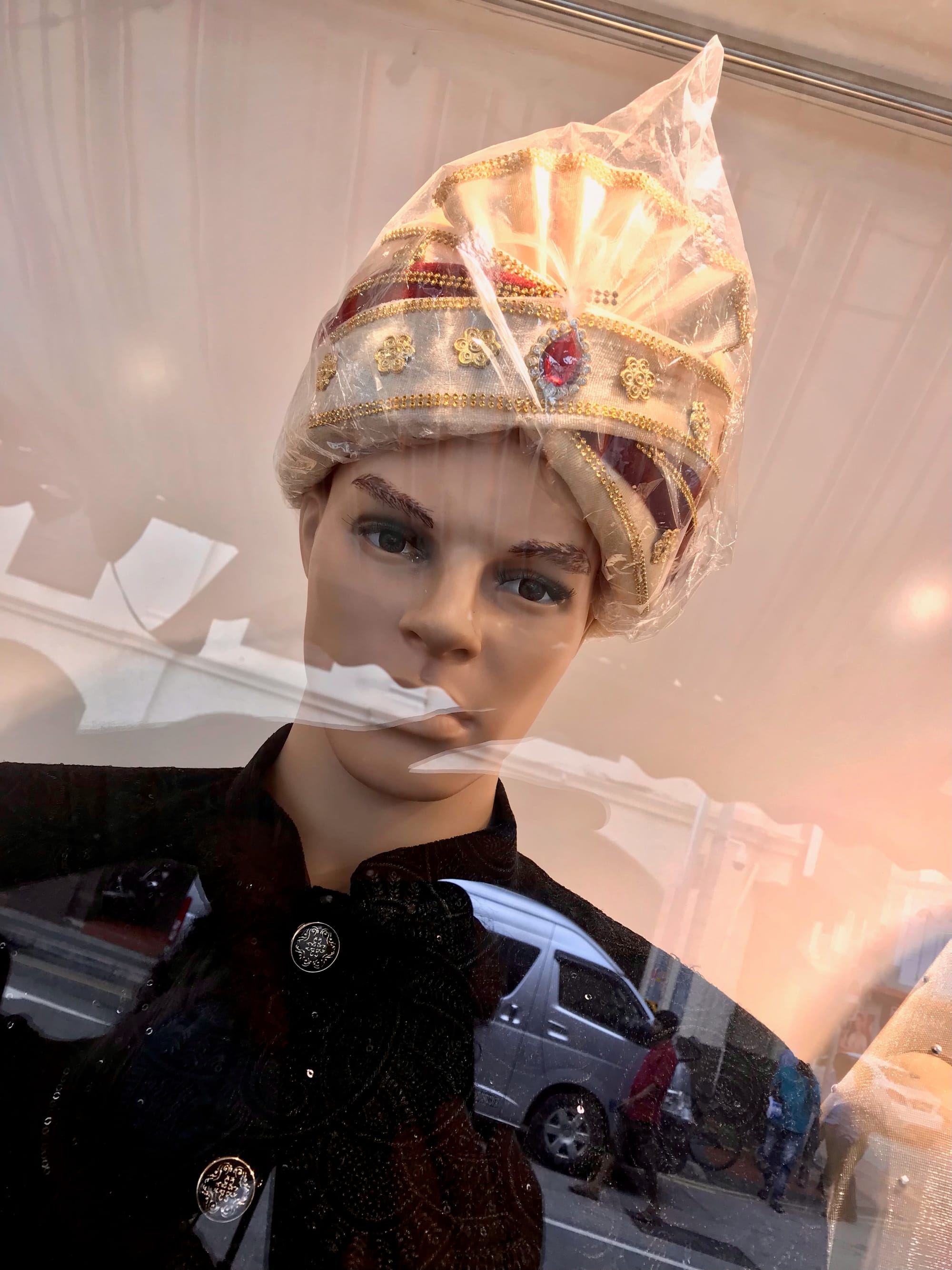 A male mannequin with a ruby-encrusted turban and navy Nehru jacket seems to look out from a glass display. A reflection of people on the street can be seen. 