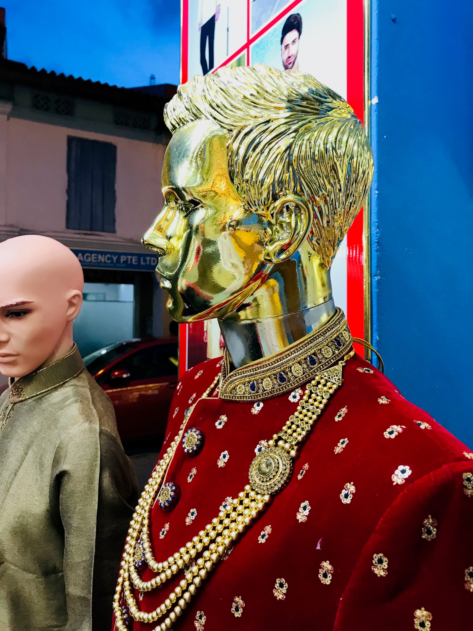 A male mannequin with a golden head wearing a red velvet jacket with a rich insignia pattern and a gold, beaded necklace.