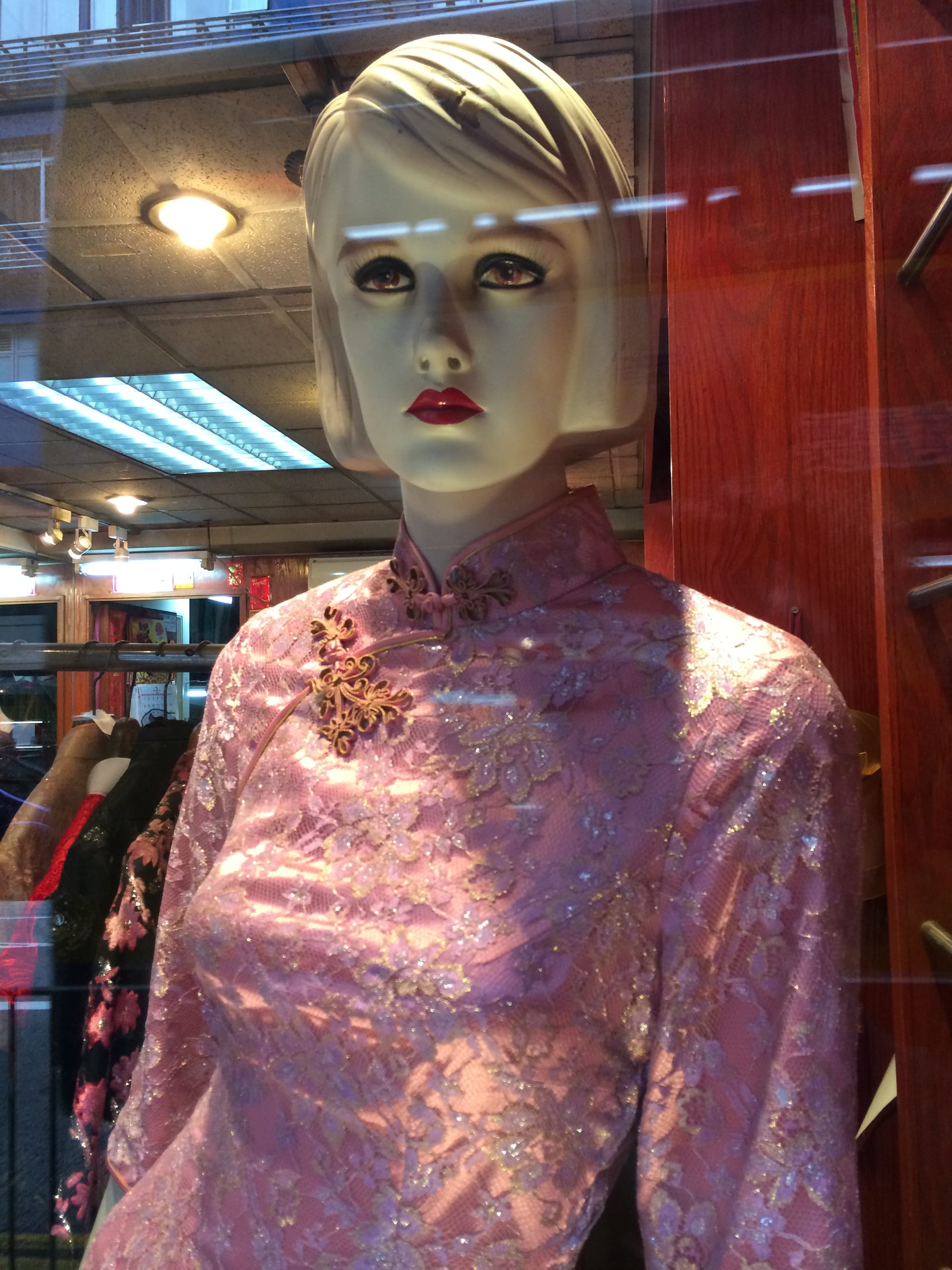 A female mannequin wearing a pink cheongsam with floral embroidery. In the glass reflection, horizontal lines of light appear above her brow.
