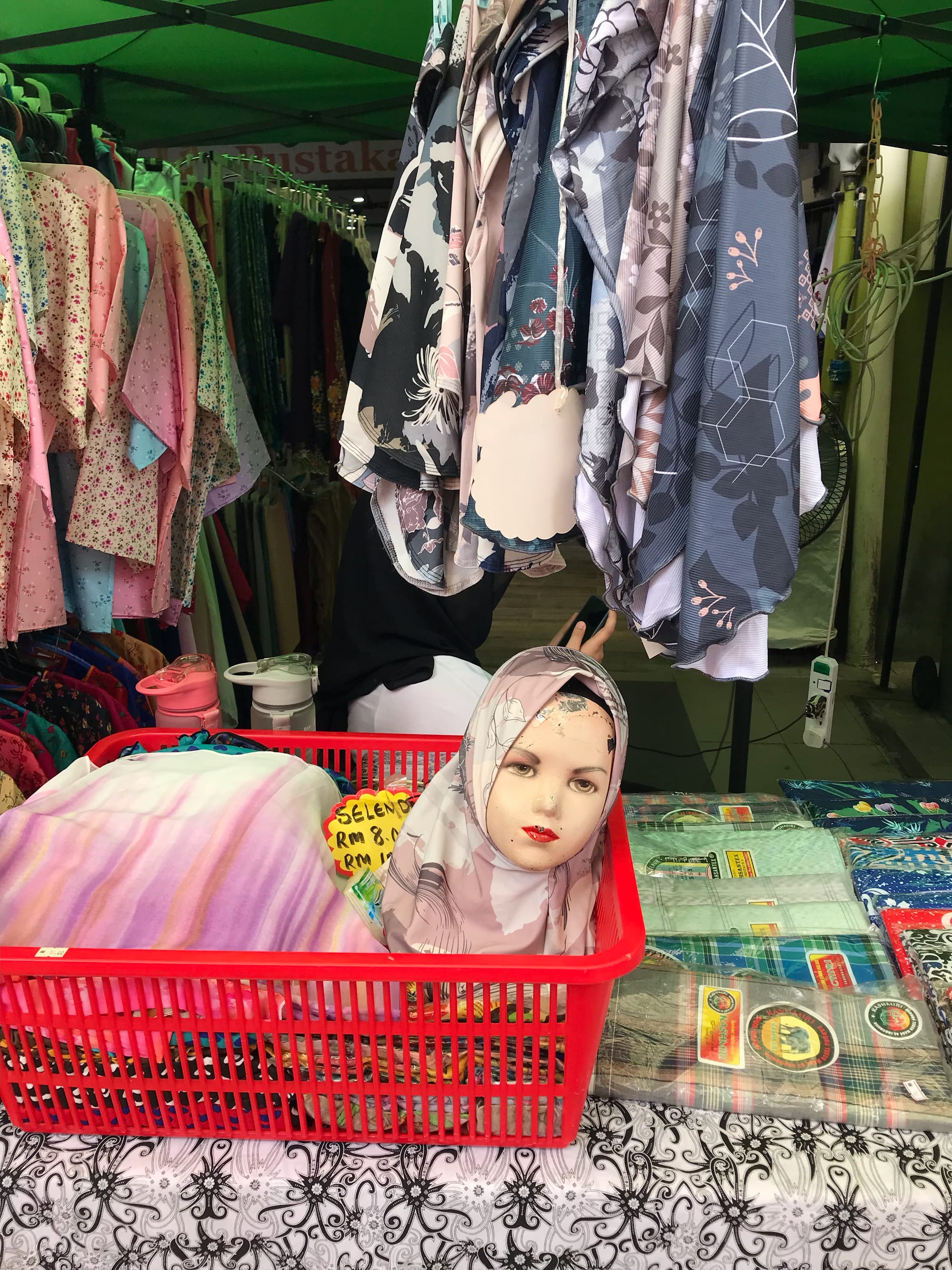 A female mannequin head, worn and cracked at the top of the forehead, wearing a pink floral hijab sits atop folded headdresses for sale in a red plastic basket on a market vendor's table.