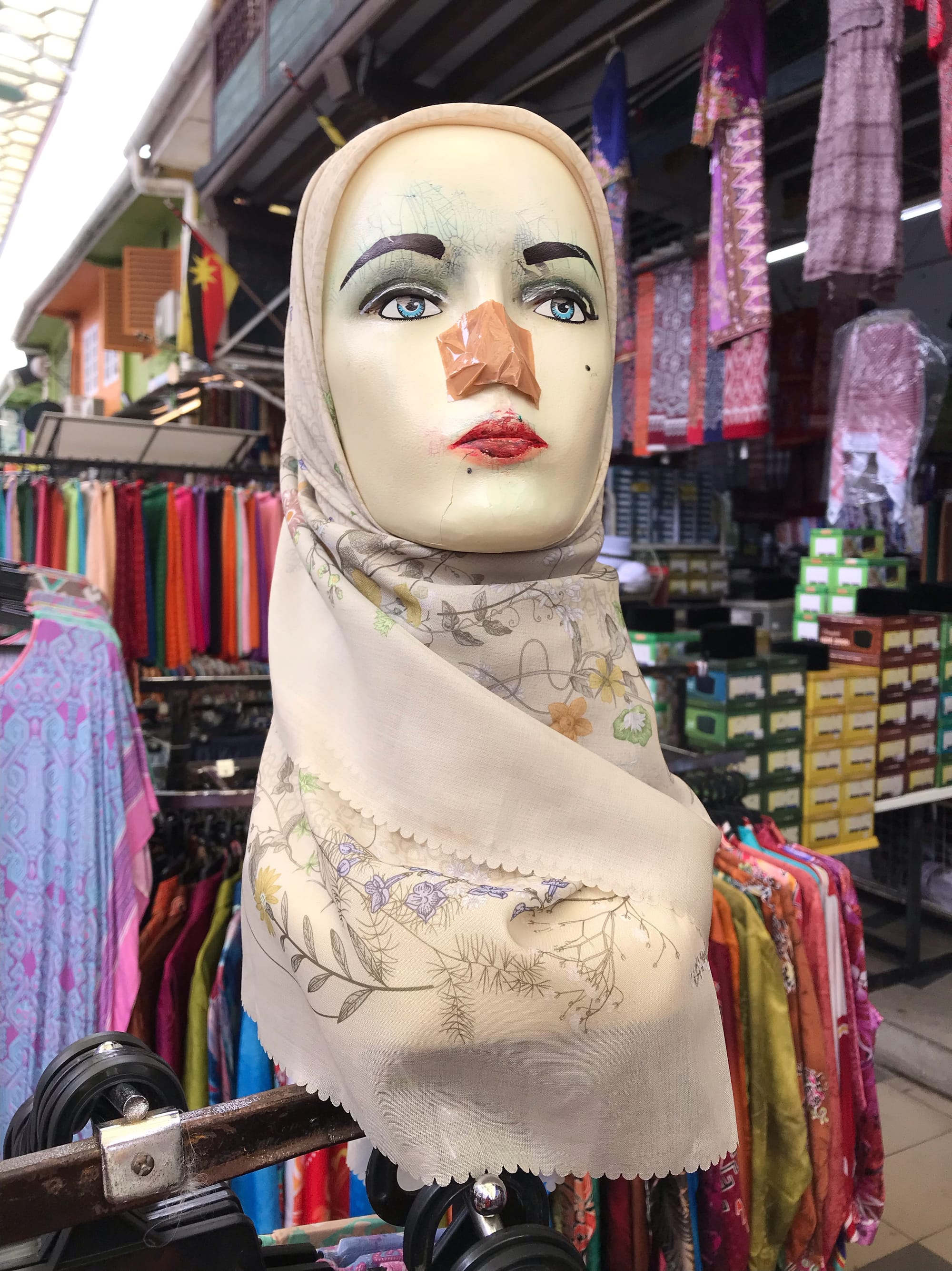 A female mannequin wears a beige hijab. The nose, perhaps missing, has been taped over with tan packing tape, the lips are scratched and messily covered in red paint. Bluish cracks appear above the eyebrows. 