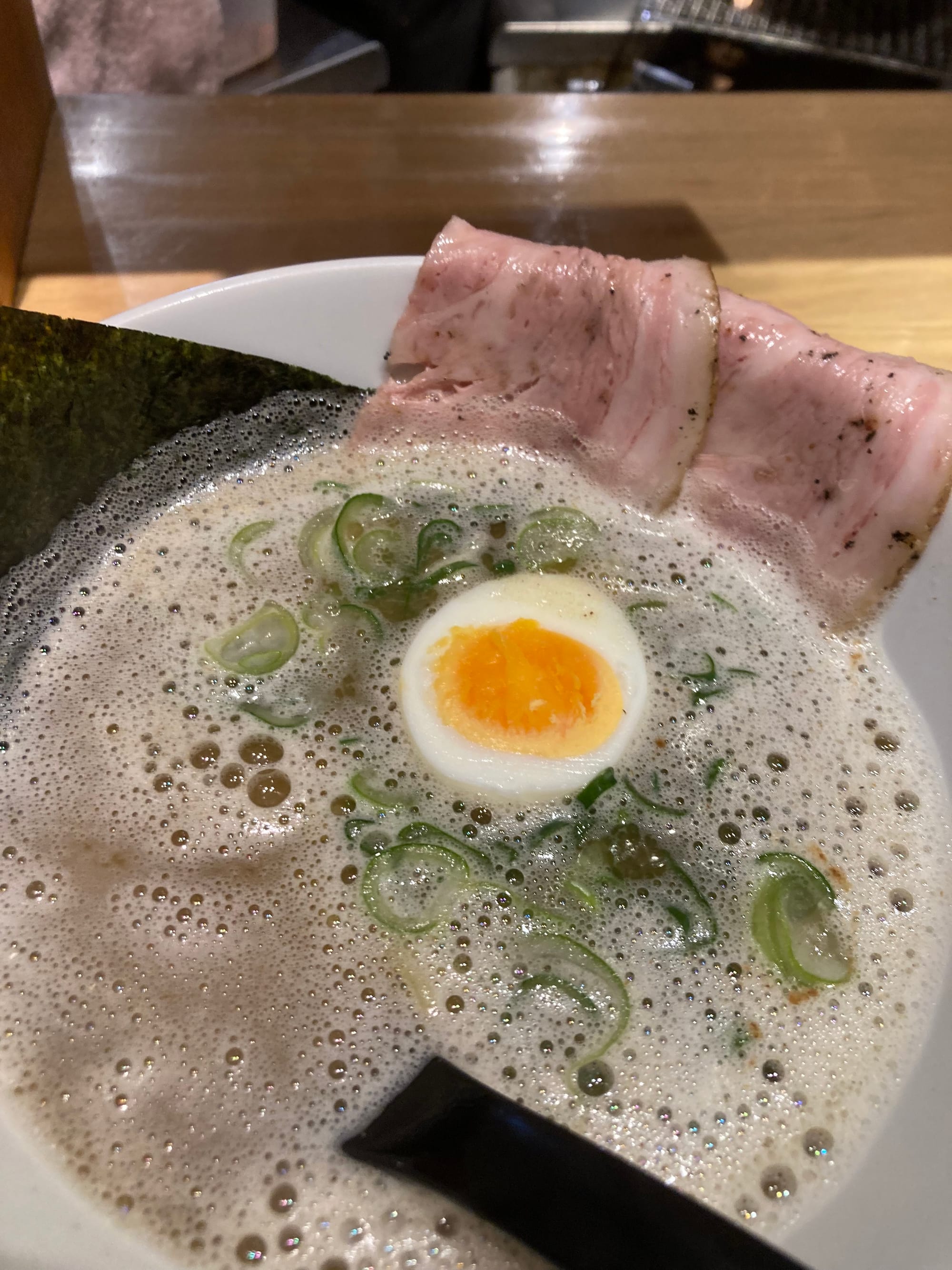 A close up of a bowl of frothy broth in a bowl. There is half a hard-boiled egg and slices of pork.