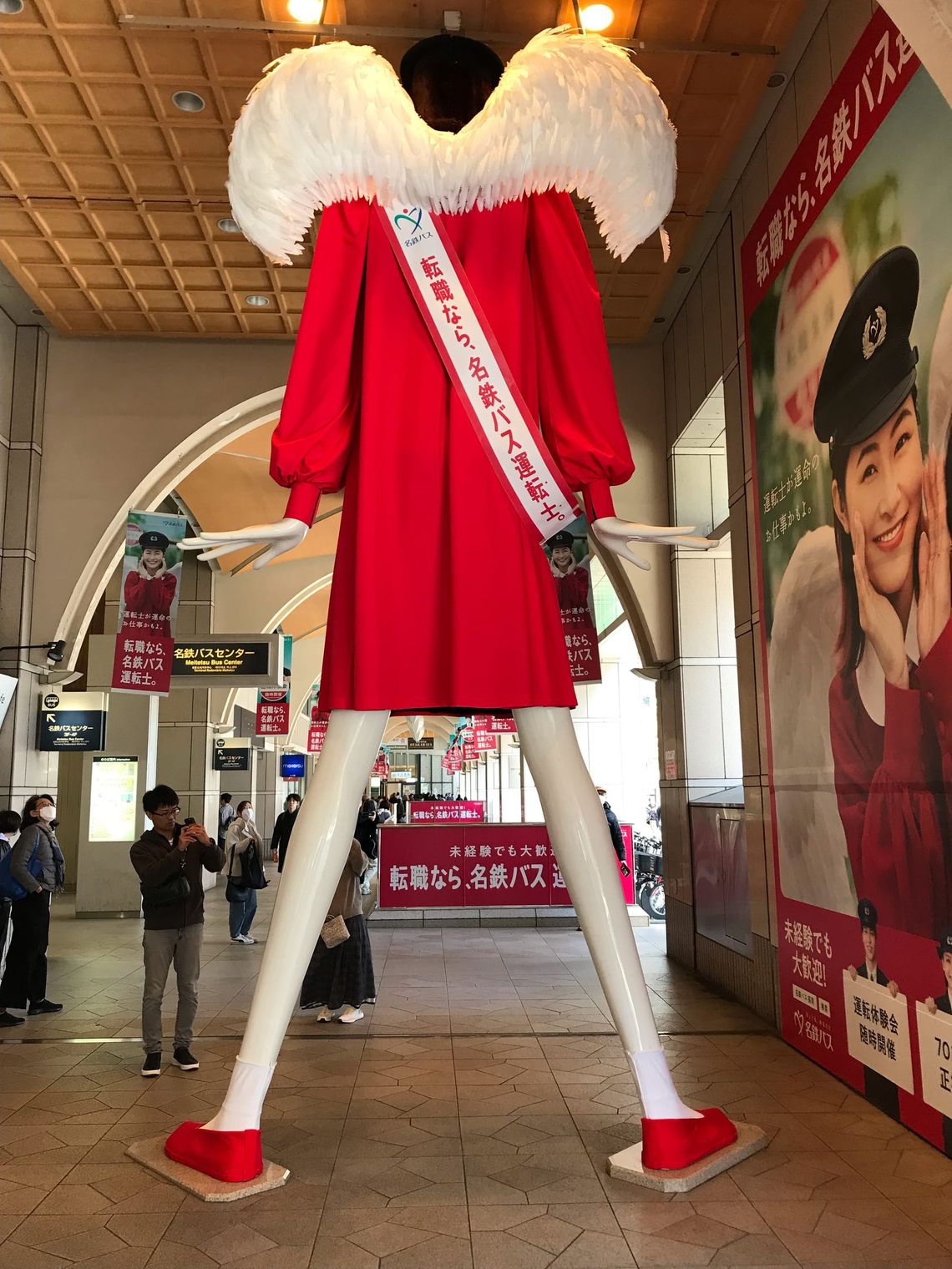 A tall white mannequin in a red dress towers over people. 