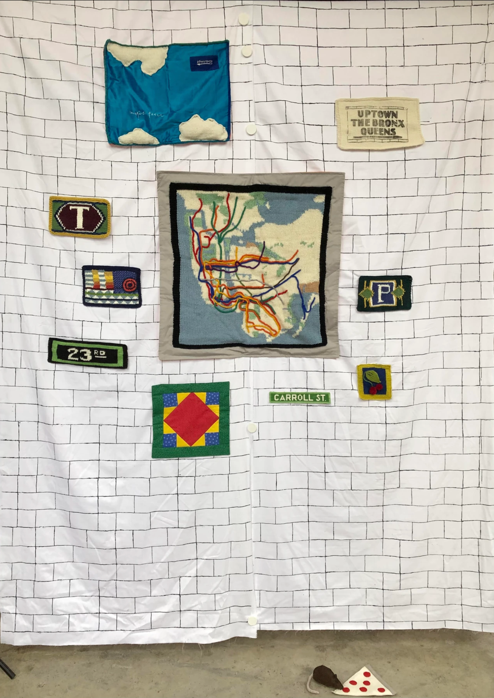 A subway wall recreation with everything made out of fiber arts including pizza rat, a subway may, and station signs