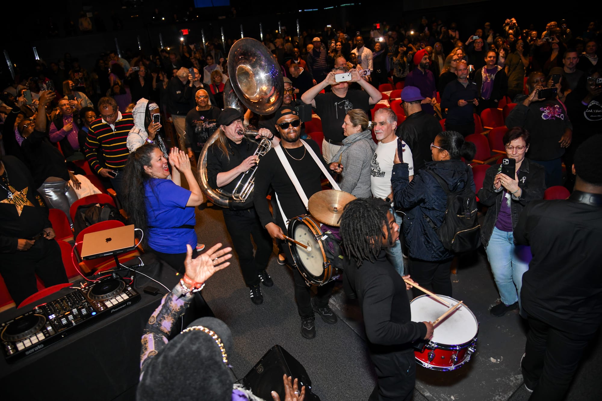 Musicians playing drums and a horns walk through a cheering crowd in the auditorium of Symphony Space
