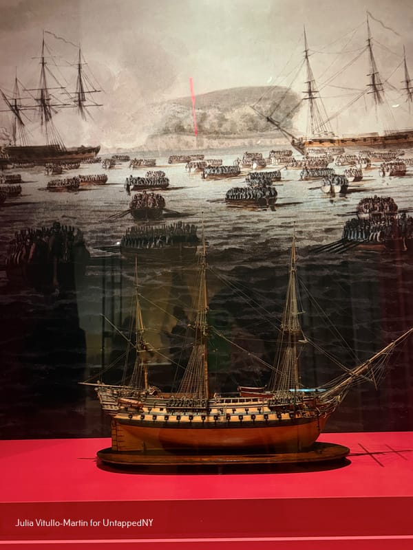 A detailed ship model displayed in front of a large historical painting depicting the British amphibious invasion of New York, with dozens of rowboats carrying troops across the water.