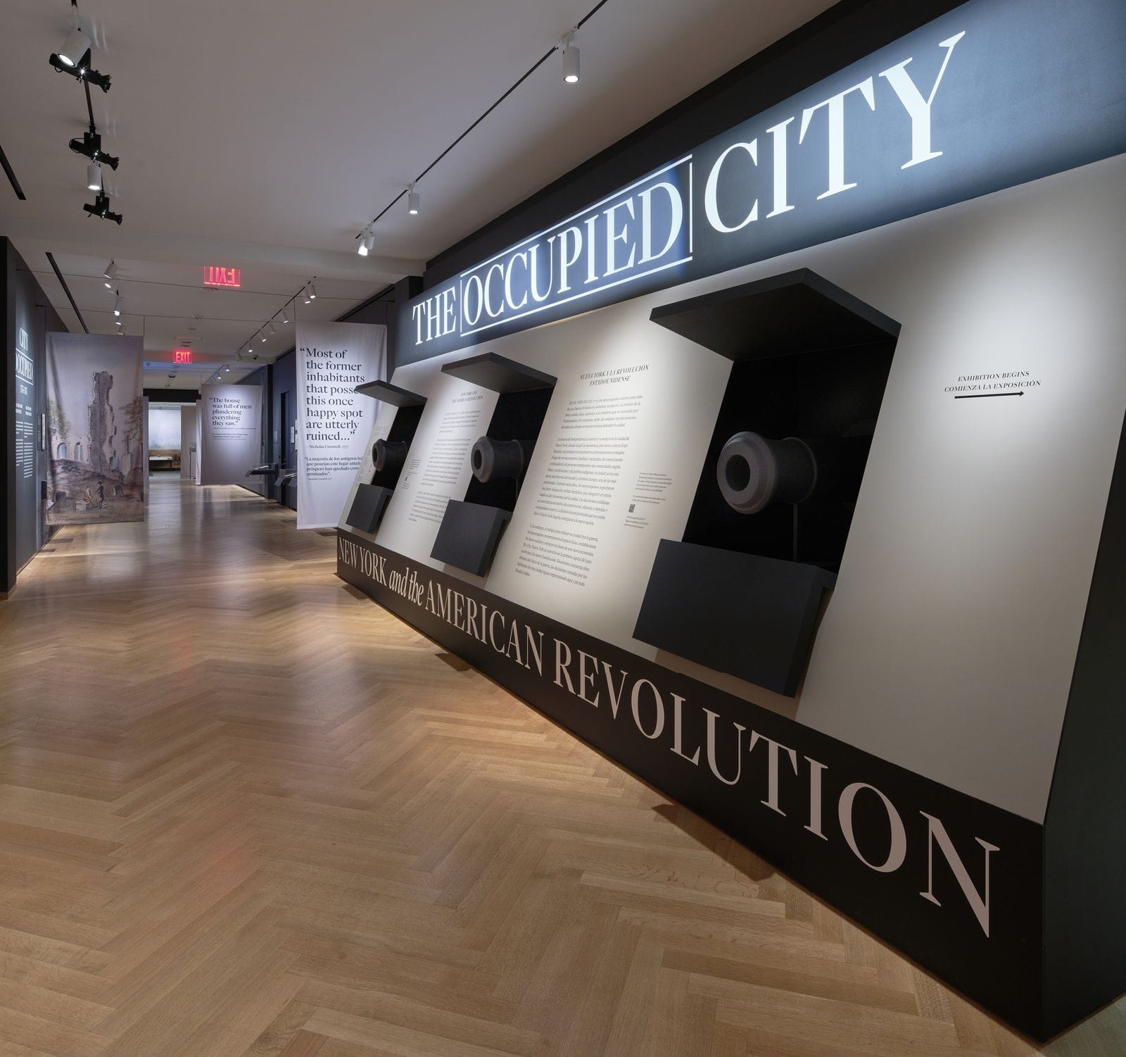 The entrance corridor of The Occupied City: New York and the American Revolution at MCNY, with the exhibit title in bold illuminated lettering above a row of mounted cannons and interpretive panels, leading into the darkened gallery beyond.
