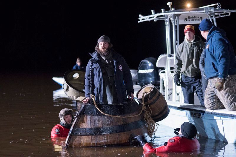 A crew member stands atop a wooden barrel-shaped vessel on the water at night during the filming of TURN: Washington's Spies, recreating the Turtle submarine used in New York Harbor during the Revolutionary War.
