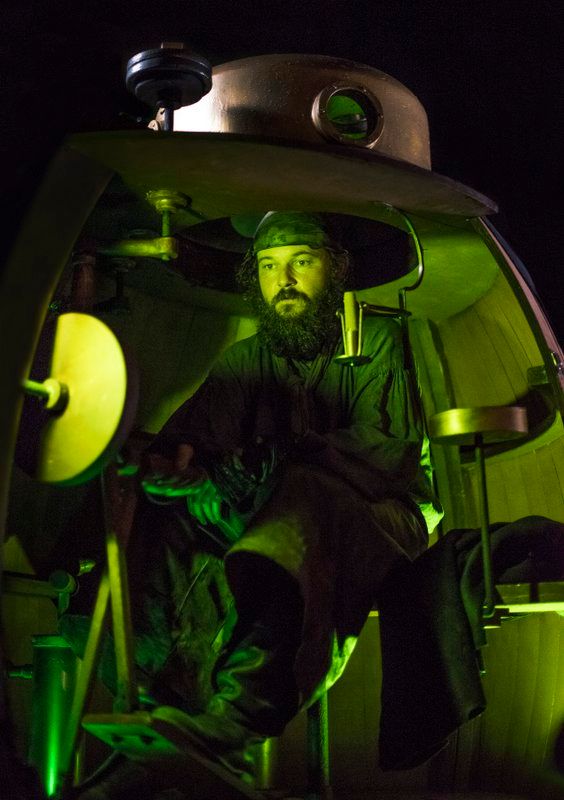 An actor seated inside a replica of the Turtle submarine, lit in eerie green, operating the hand-cranked controls of history's first combat submarine — built by David Bushnell in 1775.