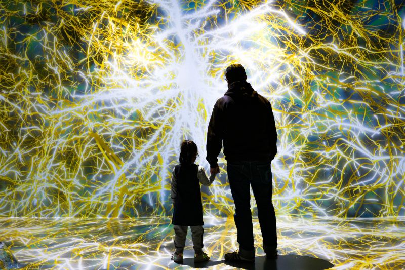An adult holds a young person's hand inside of a digital exhibition with white and yellow elements crossing the image.