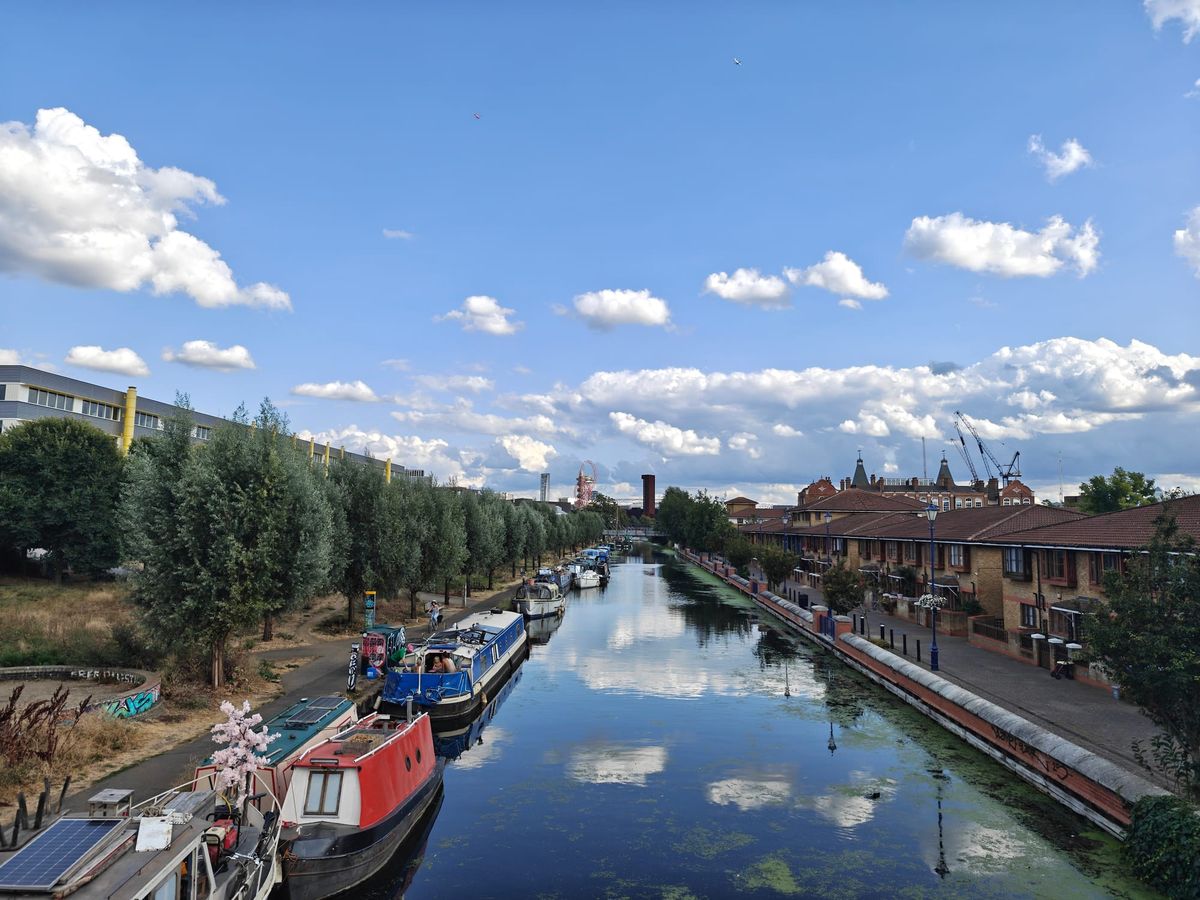 A photo taken of a canal in London, looking towards the Olympic Village; the sky is blue with little fluffy clouds peppering it; the left bank has trees adorning it, the right bank has houses