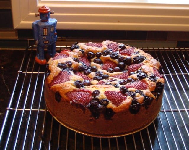 A berry-topped coffee cake on a cooling rack next to a small ceramic toy robot