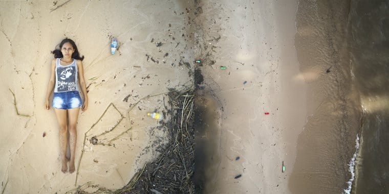 Joane deitada na areia ao lado do rio cheio de lixo plástico