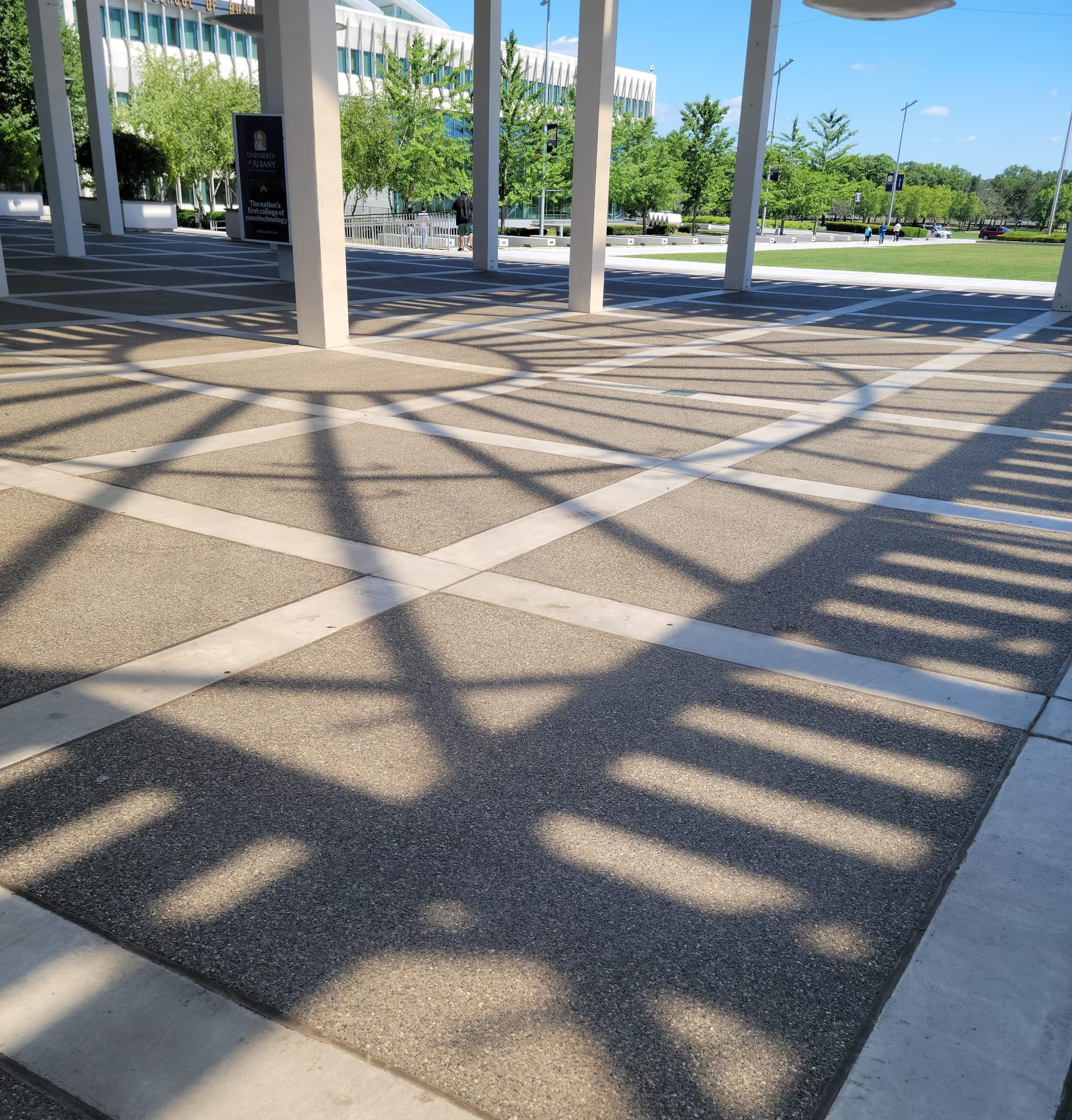 A photo of an intricate pattern of shadows projected onto a concrete plaza on the campus of UAlbany