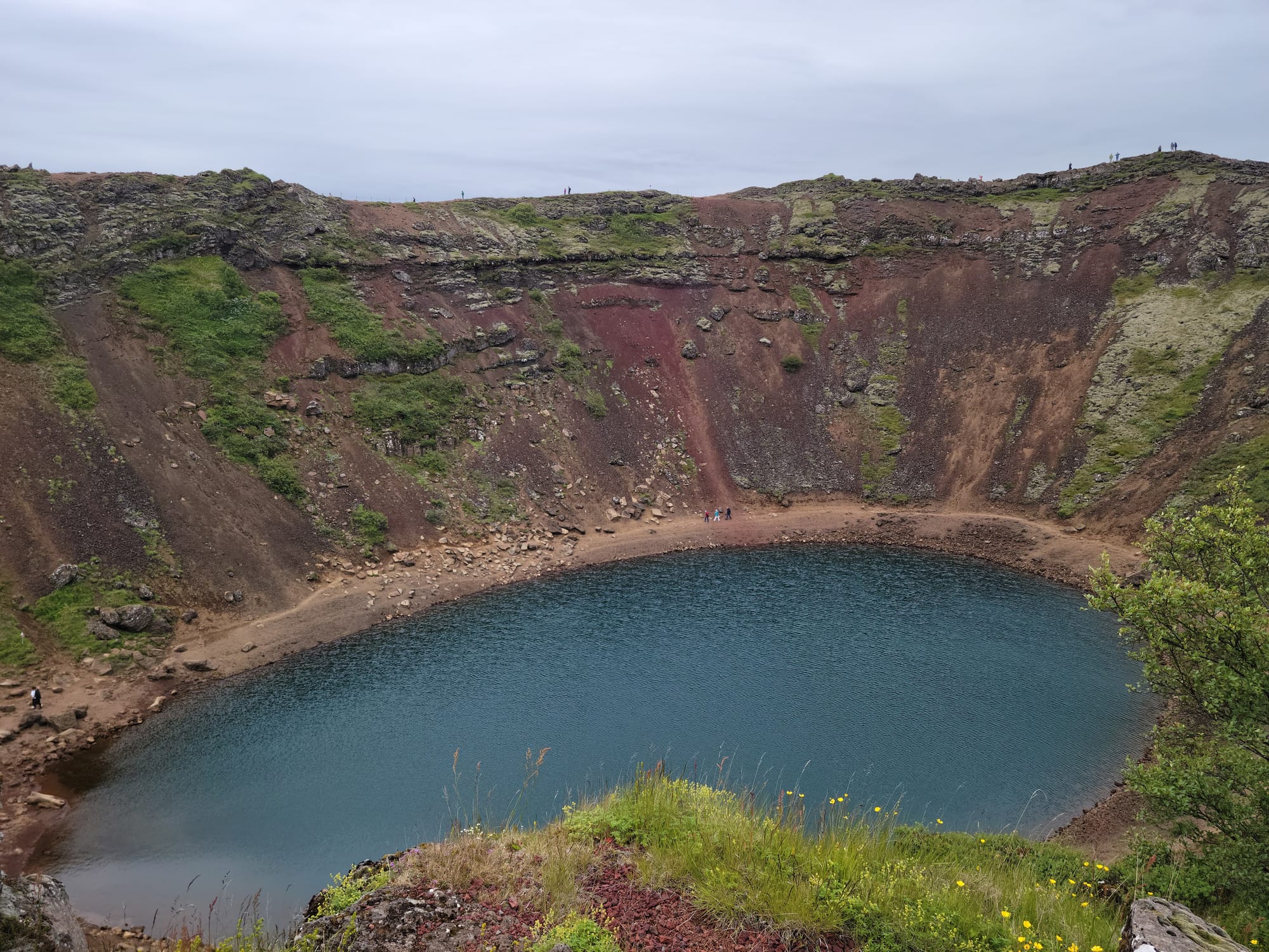 A false crater with a large dark pool at the bottom. The sides of the crater are streaked by iron deposits and covered in mossy rocks and scrub plants.