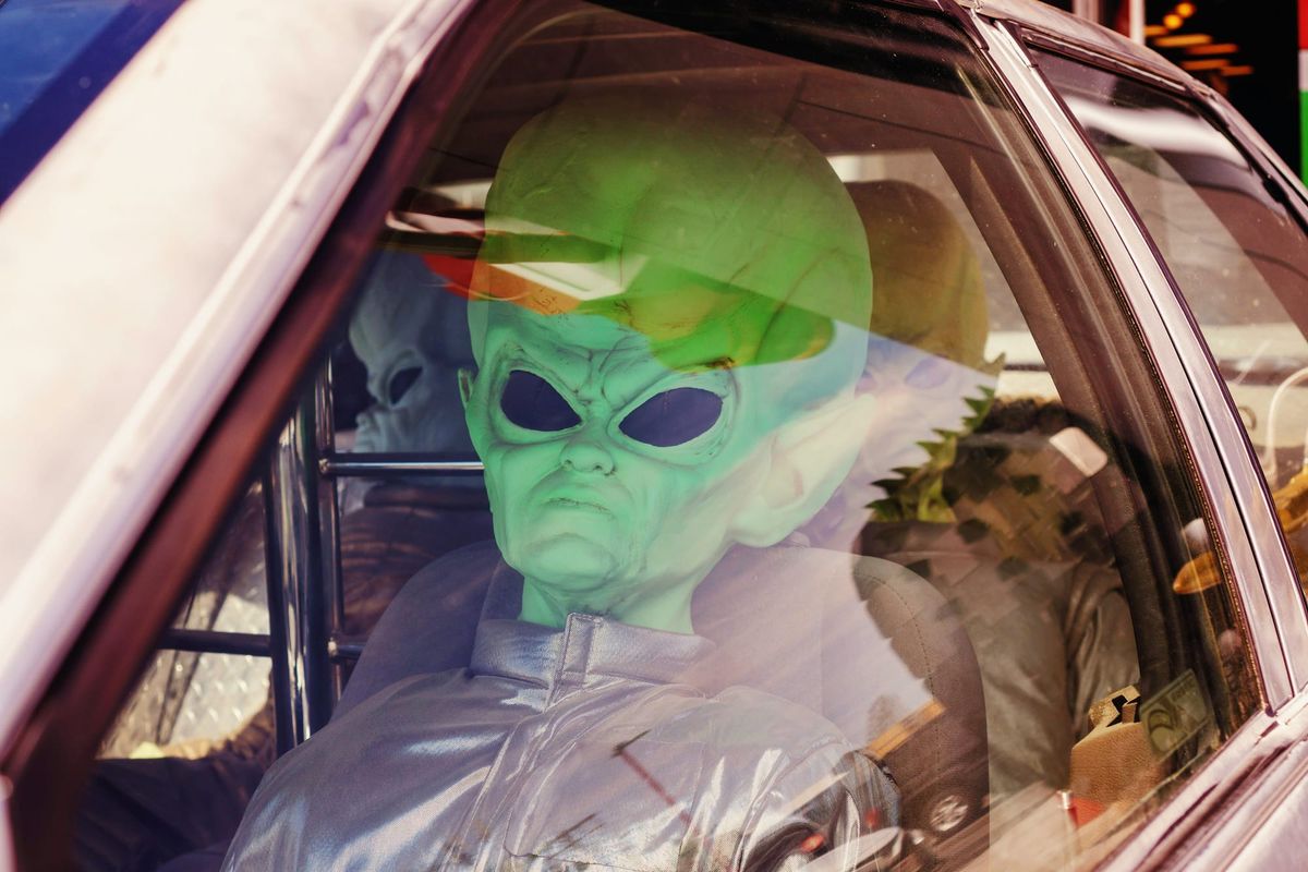A photograph of a green rubber alien in a silver jumpsuit sitting in the driver's seat of a car. Two more aliens sit in the back seat.