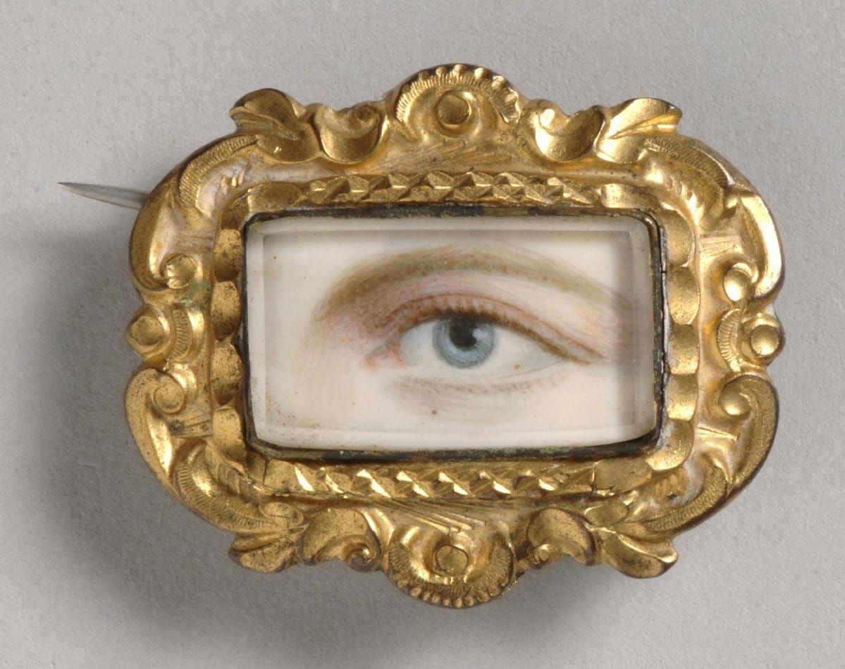 A photograph of a cameo brooch containing a drawing of a light-skinned blonde woman's blue eye and eyebrow above it.