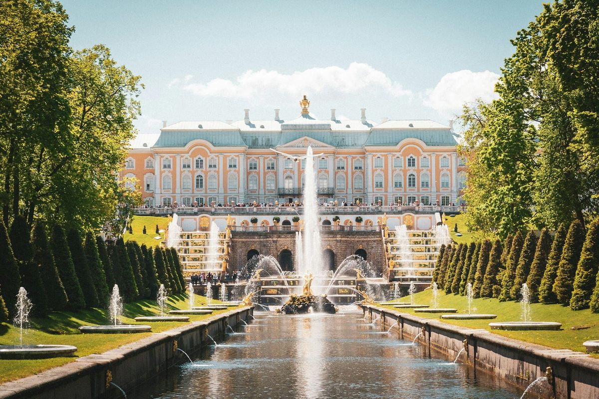 A sunny day on the fountain-filled lawn of the beautiful Peterhof Estate in St. Petersburg, Russia. At the top of the lawn sits a sprawling, well-appointed manor house.