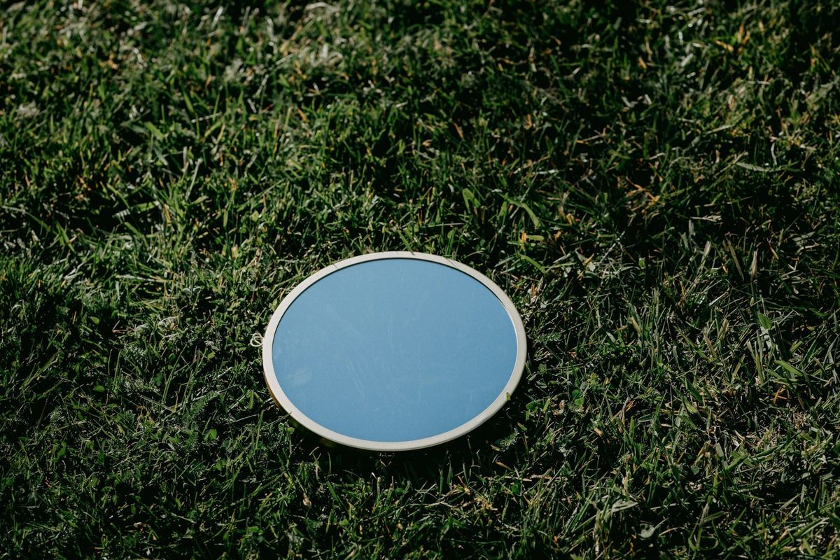 Photograph of an oval-shaped mirror sitting in the middle of a grassy field. It reflects only the blue of the sky.
