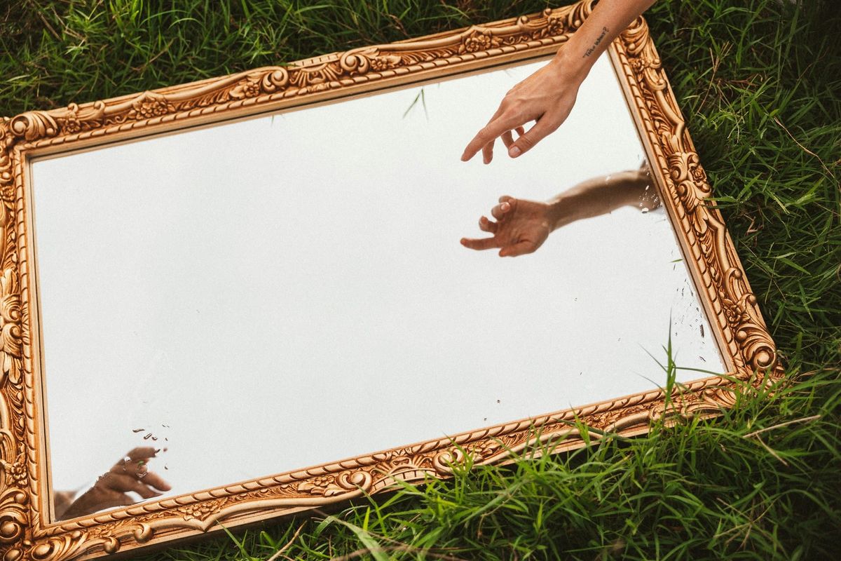 A photograph of a mirror lying in a field of grass. A hand reaches towards its surface from out of frame.