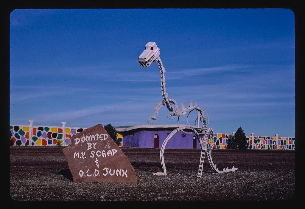 A vintage photograph of a metal statue of a dinosaur made from various forms of scrap. The dinosaur statue stands in the middle of an empty field, in front of a clear sky.