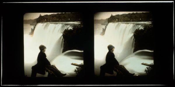 A grayscale stereograph slide of a person sitting in front of a large waterfall