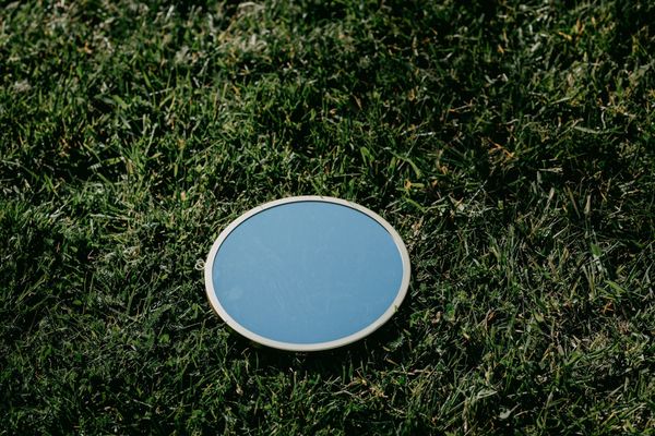 Photograph of an oval-shaped mirror sitting in the middle of a grassy field. It reflects only the blue of the sky.
