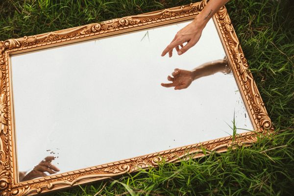 A photograph of a mirror lying in a field of grass. A hand reaches towards its surface from out of frame.
