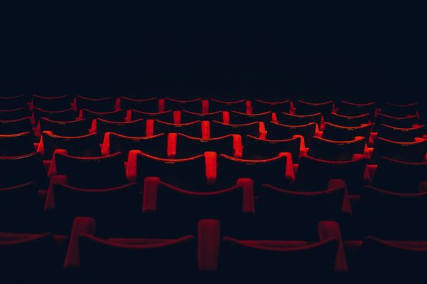A photograph of red velvet theater seating descending in rows into shadowed darkness.