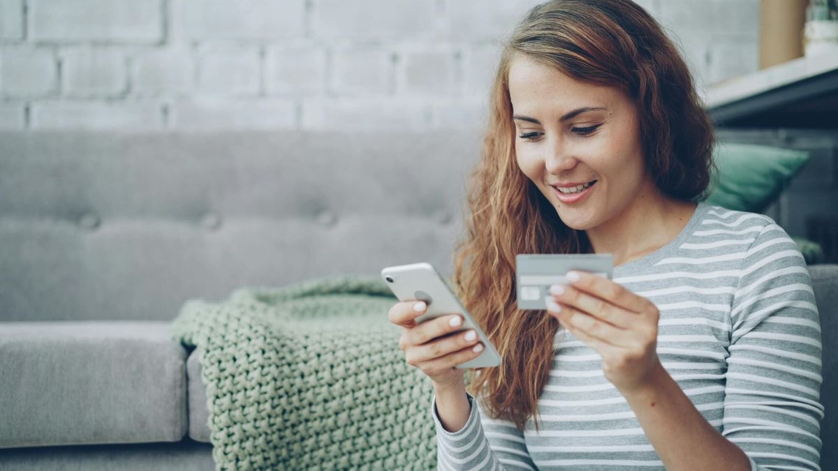 A woman with long wavy hair wearing a striped shirt holds a smartphone in one hand and a credit card in the other while sitting on a gray sofa.
