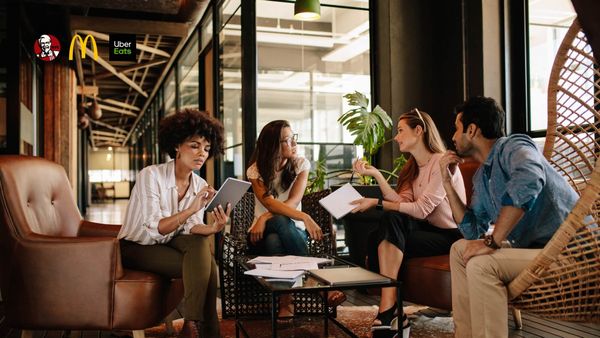 Four diverse professionals sit in a modern office lounge area, discussing work with a tablet, notebooks, and papers on a coffee table between them.