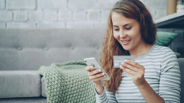 A woman with long wavy hair wearing a striped shirt holds a smartphone in one hand and a credit card in the other while sitting on a gray sofa.