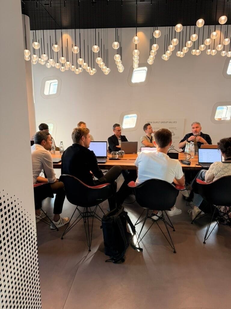 Participants with laptops around a long table under pendant lights during the AI workshop on day two.
