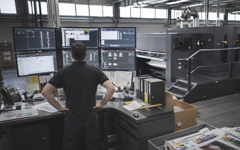 A print operator at a control desk with several screens, next to a press on the production floor.