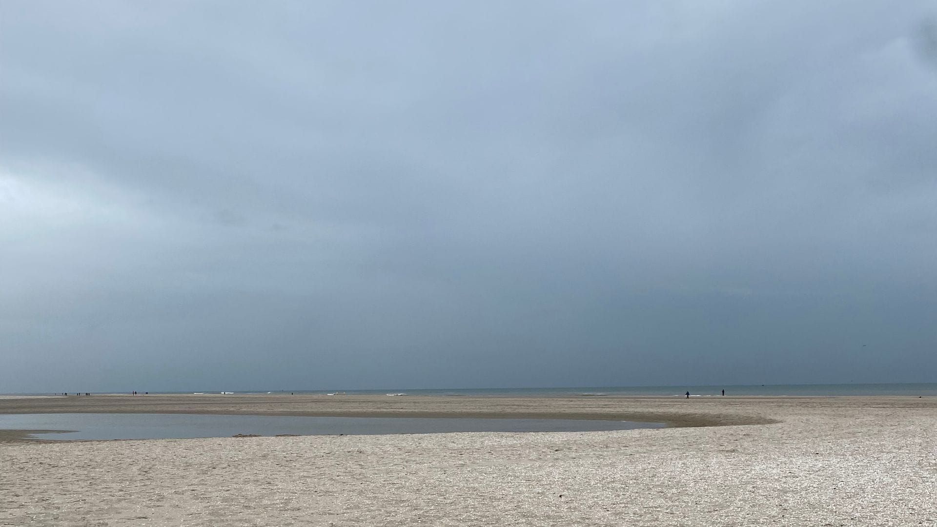Een afbeelding van een strand met op de voorgrond een zeebank en aan de horizon zie je de zee en schimmen van mensen onder een bewolkte lucht.
