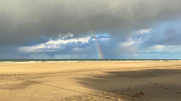 Een verlaten breed zandstrand waarop schaduwen en de warmte van de zon zichtbaar zijn met daarboven een donkere dreigende wolken en in de verte een regenboog.