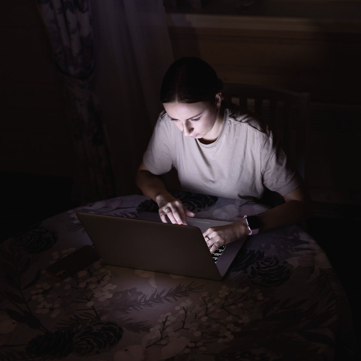 Woman in white shirt using laptop on bed in dark room at night