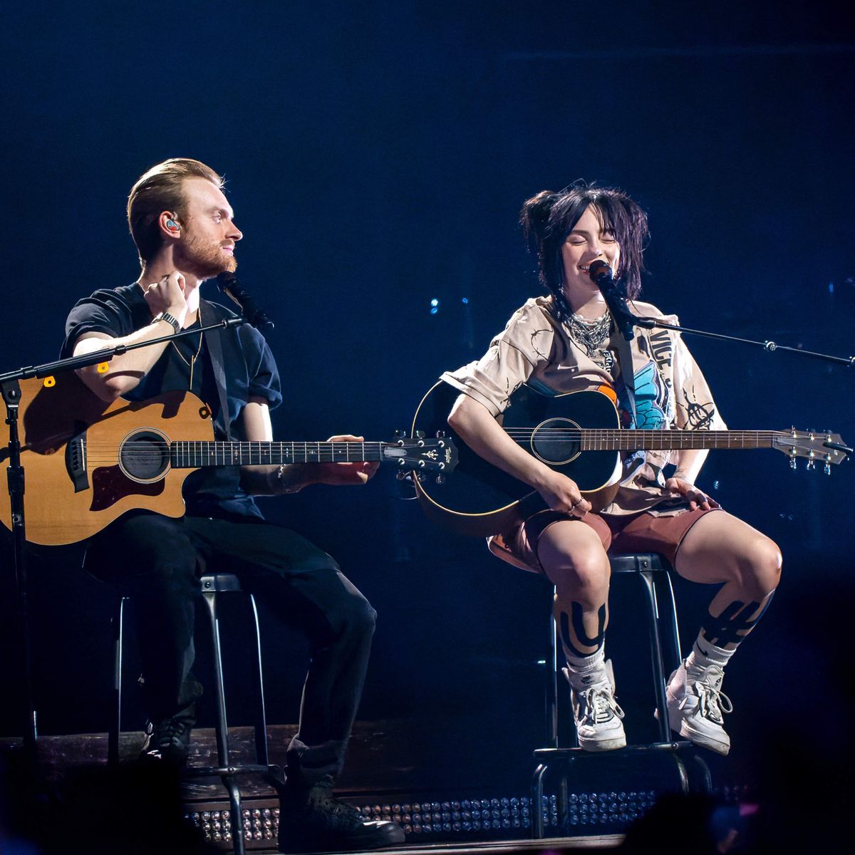 Two musicians performing on stage, playing acoustic guitars while seated on stools