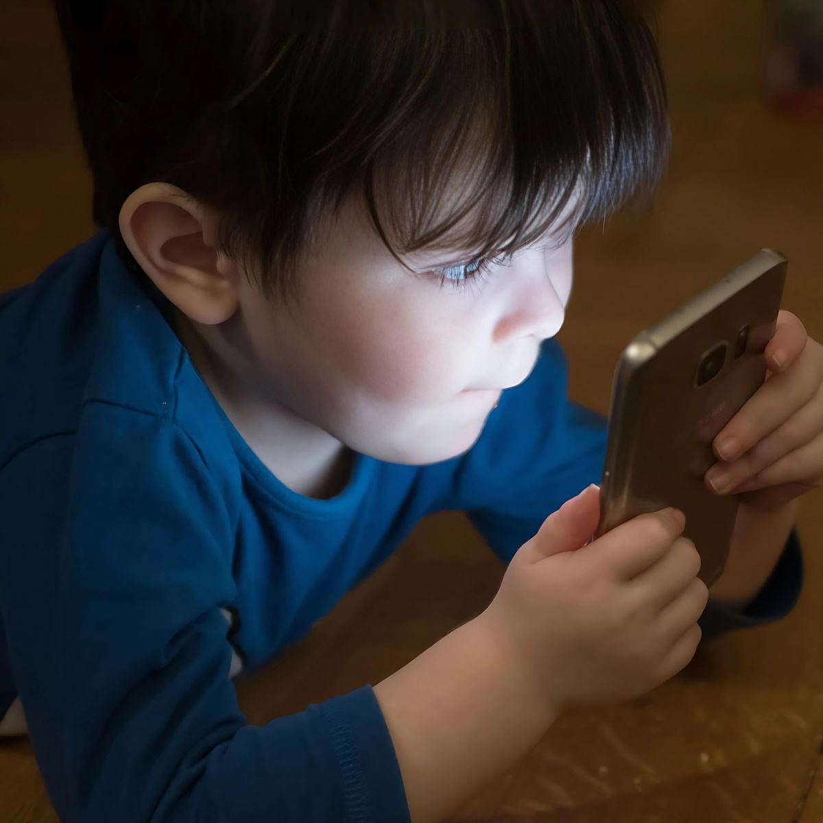 Young boy in blue shirt intensely focused on smartphone screen in dim lighting