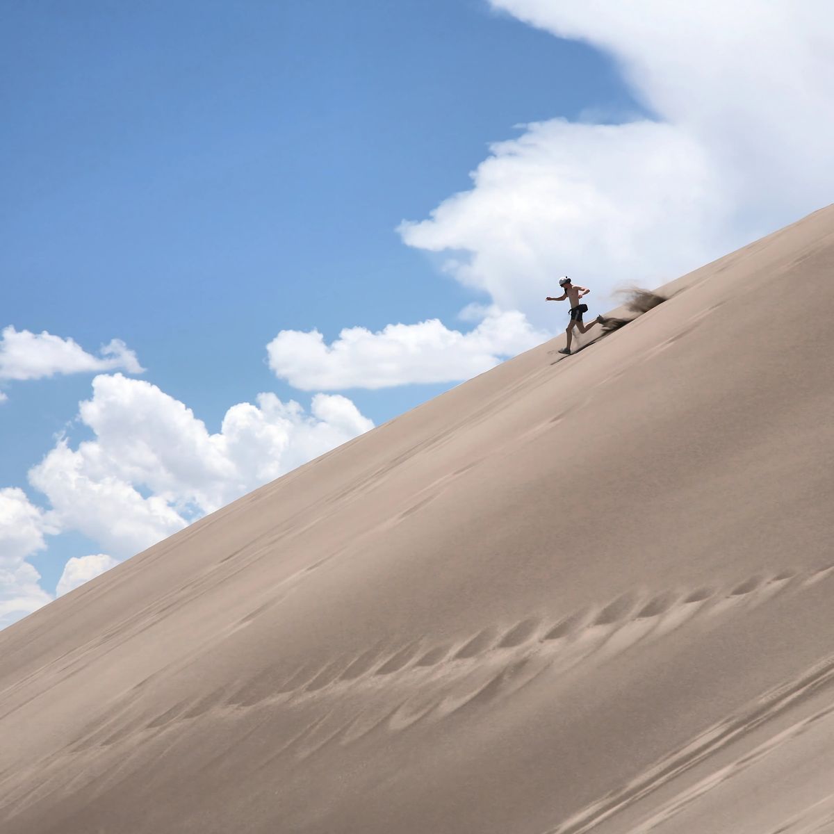 Person sandboarding down steep dune under bright blue sky with scattered white clouds