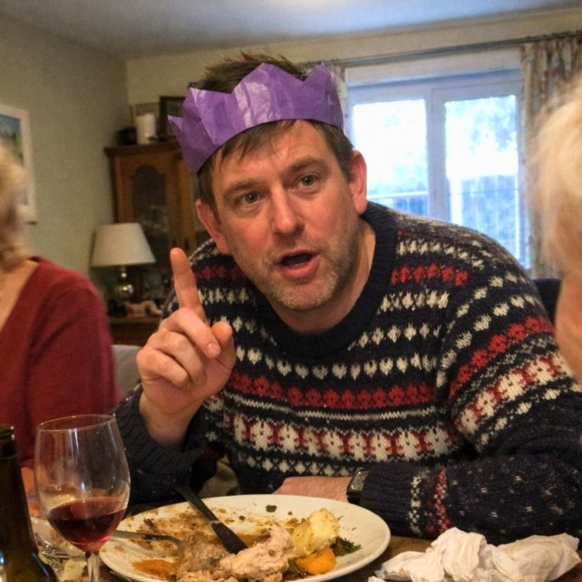 Man in paper crown raising finger mid-lecture at Christmas dinner table