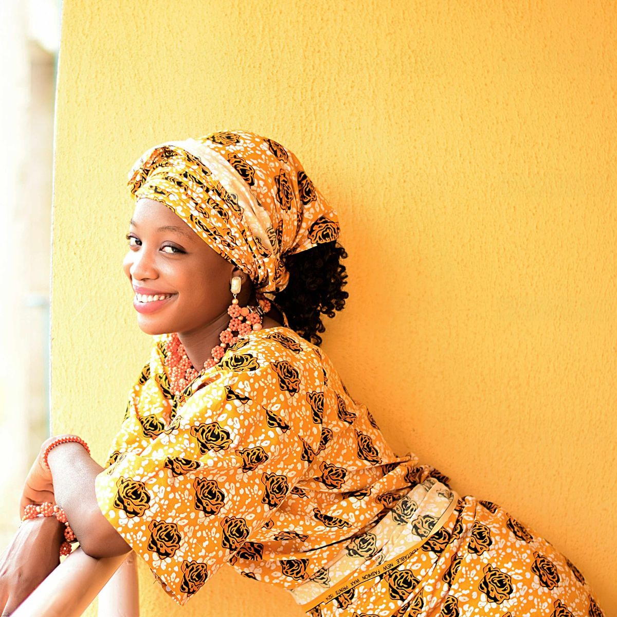 Woman in orange patterned outfit smiling while leaning against yellow wall