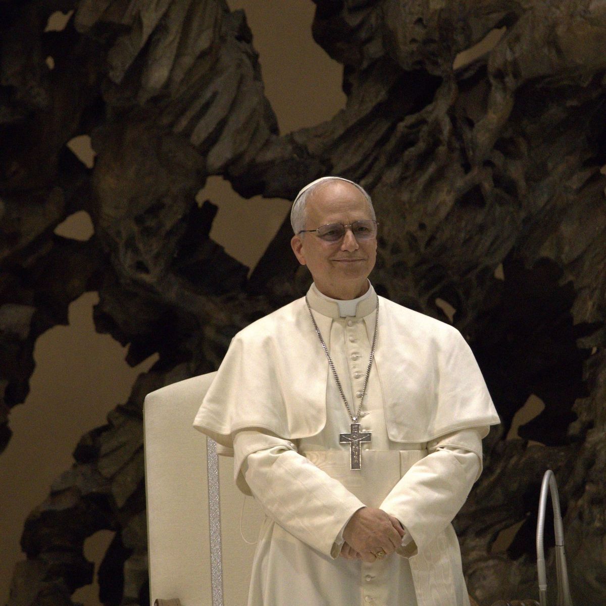 A pope in white robes stands smiling before a dark sculpture