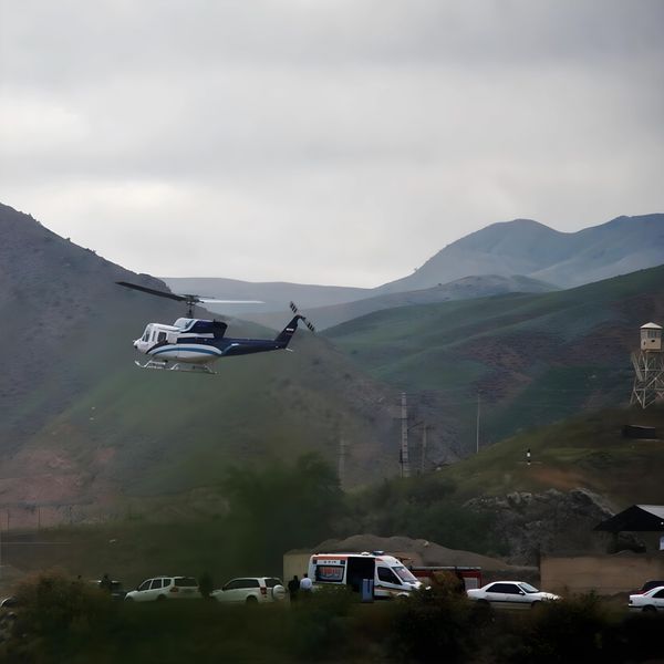 Helicopter flying over mountainous terrain with emergency vehicles and watchtower visible below