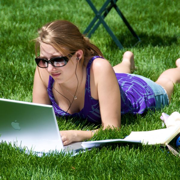 Young woman lying on grass, using laptop and wearing headphones and sunglasses