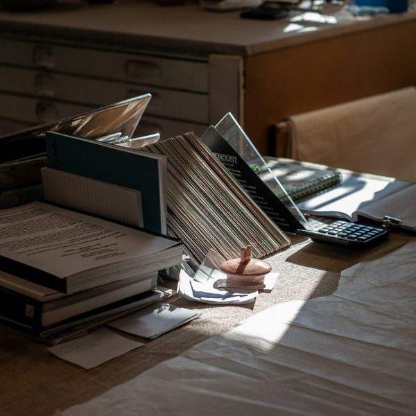 Cluttered desk with stacked documents, folders, and a calculator in dim afternoon light, chair empty