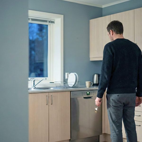 Man in sweater stands alone in sparse kitchen holding fork, dishwasher glowing beside him